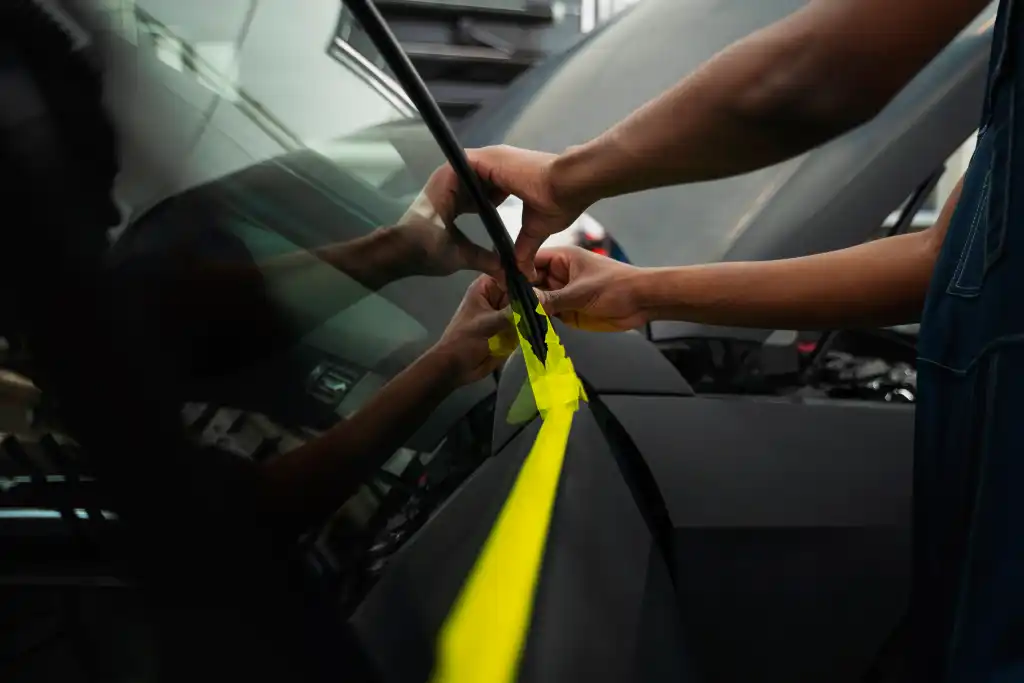 Hands applying yellow tape to secure car windscreen before tinting
